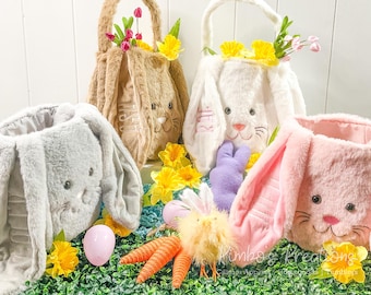 May include: Four plush bunny-shaped Easter baskets in pink, white, brown, and gray. Each basket has a fluffy bunny face with embroidered eyes and a nose. The baskets are filled with colorful flowers and a purple stuffed bunny.