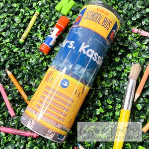 May include: A yellow school bus tumbler with a blue and white design. The tumbler has the text "School Bus" and "Mrs. Kassie" on it.