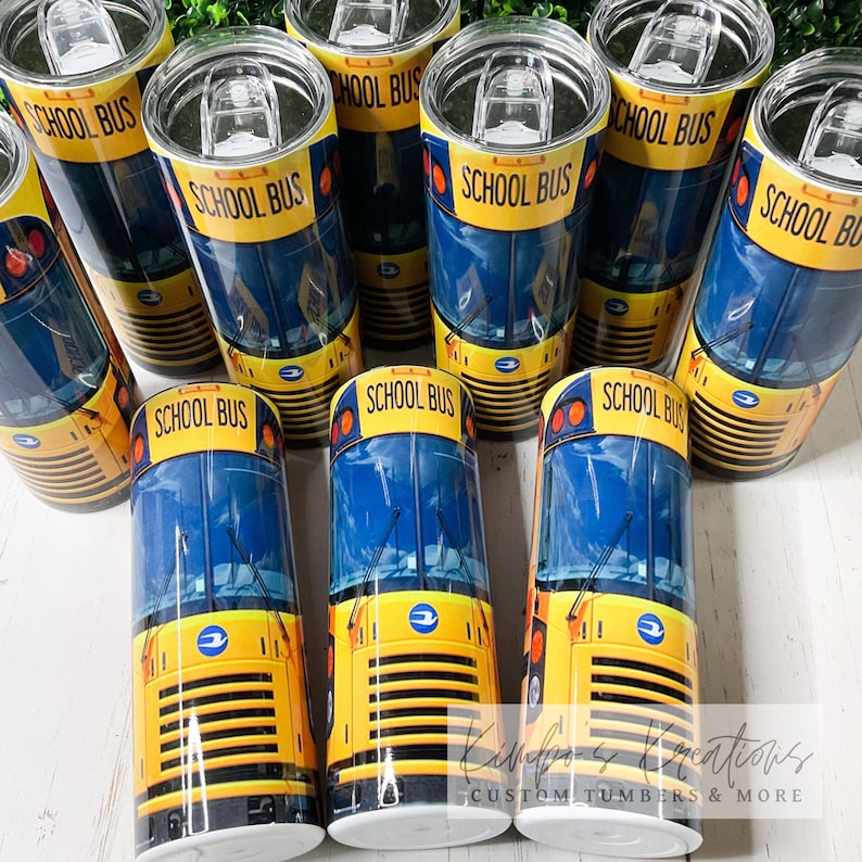 May include: A group of yellow school bus tumblers with blue windows and the words "School Bus" printed on the front. The tumblers are arranged in a semi-circle on a wooden surface.
