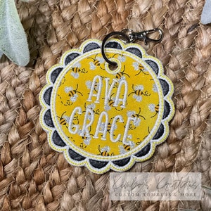 May include: A yellow and black bee-themed fabric keychain with the name "AVA GRACE" embroidered in white. The keychain is attached to a silver key ring.