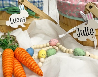 May include: Two wooden Easter baskets with bunny-shaped tags, one blue and white checkered and the other pink with floral print. The tags are labeled "Lucas" and "Julie". The baskets are surrounded by carrots and a string of wooden beads with colorful speckled eggs.