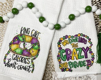 May include: Two white kitchen towels with Mardi Gras themed designs. One towel features a purple, green, and gold king cake with the text "King Cake Calories don't count". The other towel features a purple, green, and gold circle with the text "We Don't Hide The Crazy We Parade It".