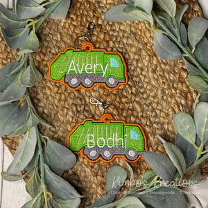 May include: Two green and orange embroidered keychain ornaments shaped like garbage trucks. One says "Avery" and the other says "Bodhi" in white thread. The keychains have silver-colored clasps. The background is a woven mat with green leaves.