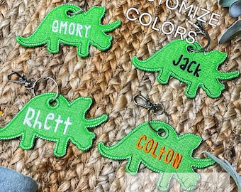 May include: Four green embroidered dinosaur-shaped keychains with different names: Emory, Jack, Rhett, and Colton. The keychains are on a woven brown background with the text "Customize Colors".