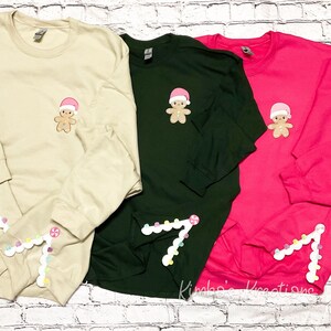 May include: Three long-sleeved shirts with a gingerbread man design on the front and a candy cane design on the sleeve. The shirts are beige, green, and pink.
