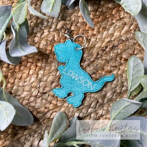 May include: A turquoise blue dinosaur-shaped keychain with the name "LAWSON" embroidered on it. The keychain is made of faux leather and has a silver metal ring and clasp.