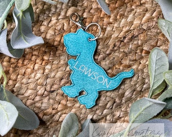 May include: A turquoise blue dinosaur-shaped keychain with the name "LAWSON" embroidered on it. The keychain is made of faux leather and has a silver metal ring and clasp.