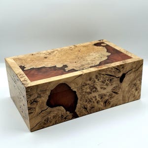 May include: A rectangular wooden box featuring a natural burl wood grain pattern with light tan and dark brown swirls. The lid and front panel incorporate smooth, reddish-brown resin inlays that contrast with the textured, knotty wood surface.
