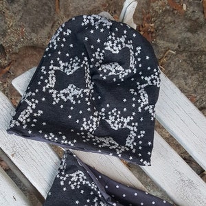 Könnte beinhalten: Ein schwarzes und weißes gemustertes Mützen- und Halssocken-Set. Mütze und Halssocken haben ein sich wiederholendes Muster aus Sternen und einem stilisierten Tierdesign.