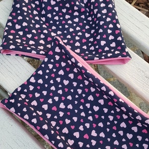 Könnte beinhalten: Eine marineblaue Mütze und ein passendes Stirnband mit einem rosa Herzmuster. Mütze und Stirnband sind aus einem weichen, dehnbaren Stoff.