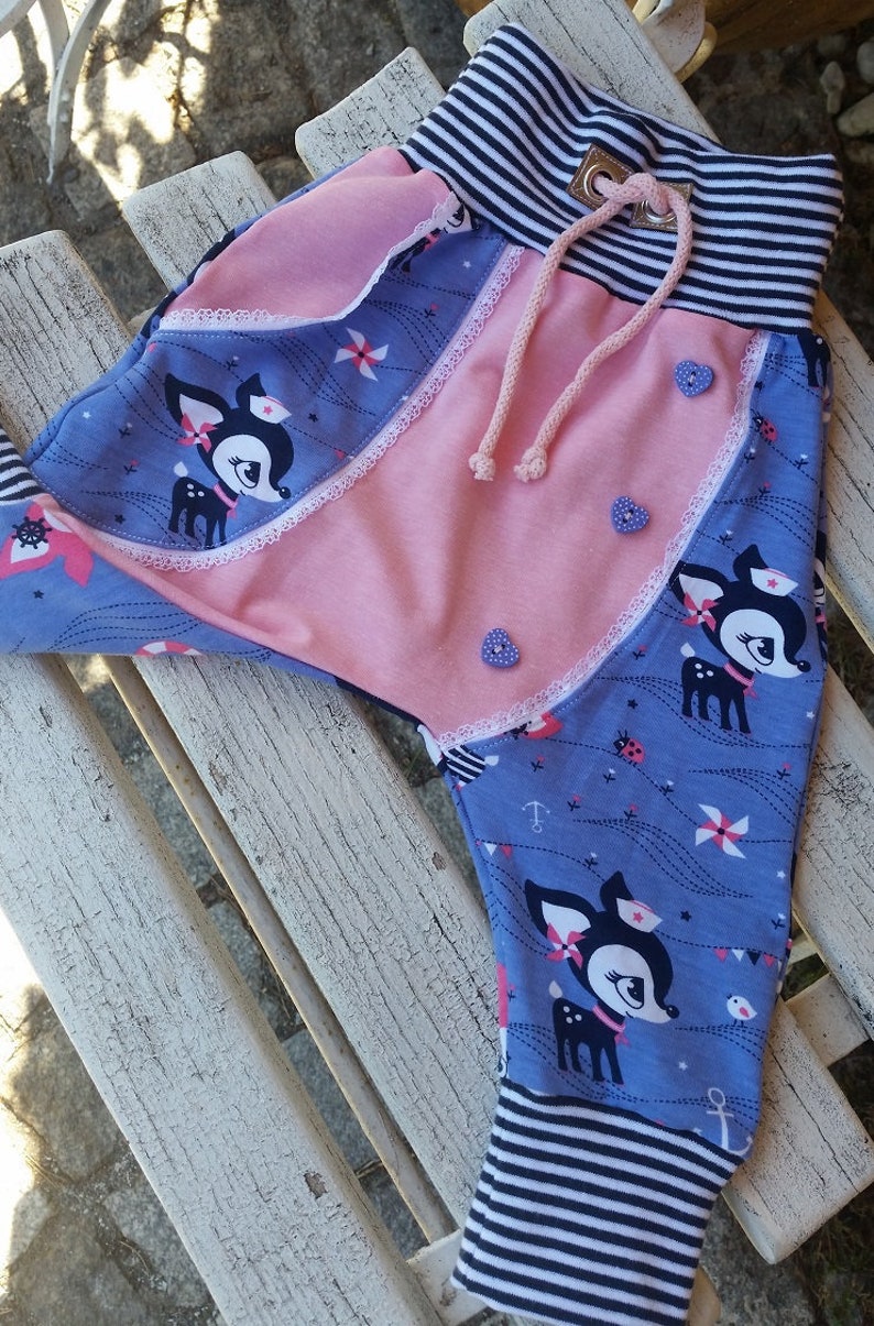 K&ouml;nnte beinhalten: Blaue und rosa Babyhose mit einem Hirschdruck und wei&szlig;em Besatz. Die Hose hat einen gestreiften Bund und einen Kordelzug.