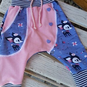 K&ouml;nnte beinhalten: Rosa und blaue Babyhose mit einem Hirschdruck und wei&szlig;er Spitzenborte. Die Hose hat einen gestreiften Bund und einen Kordelzug.