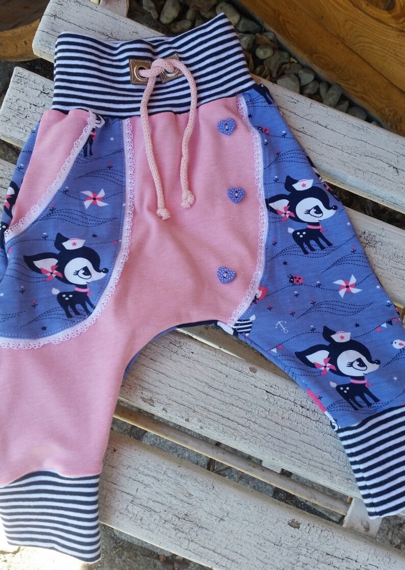 K&ouml;nnte beinhalten: Rosa und blaue Babyhose mit einem Hirschdruck und wei&szlig;er Spitzenborte. Die Hose hat einen gestreiften Bund und einen Kordelzug.