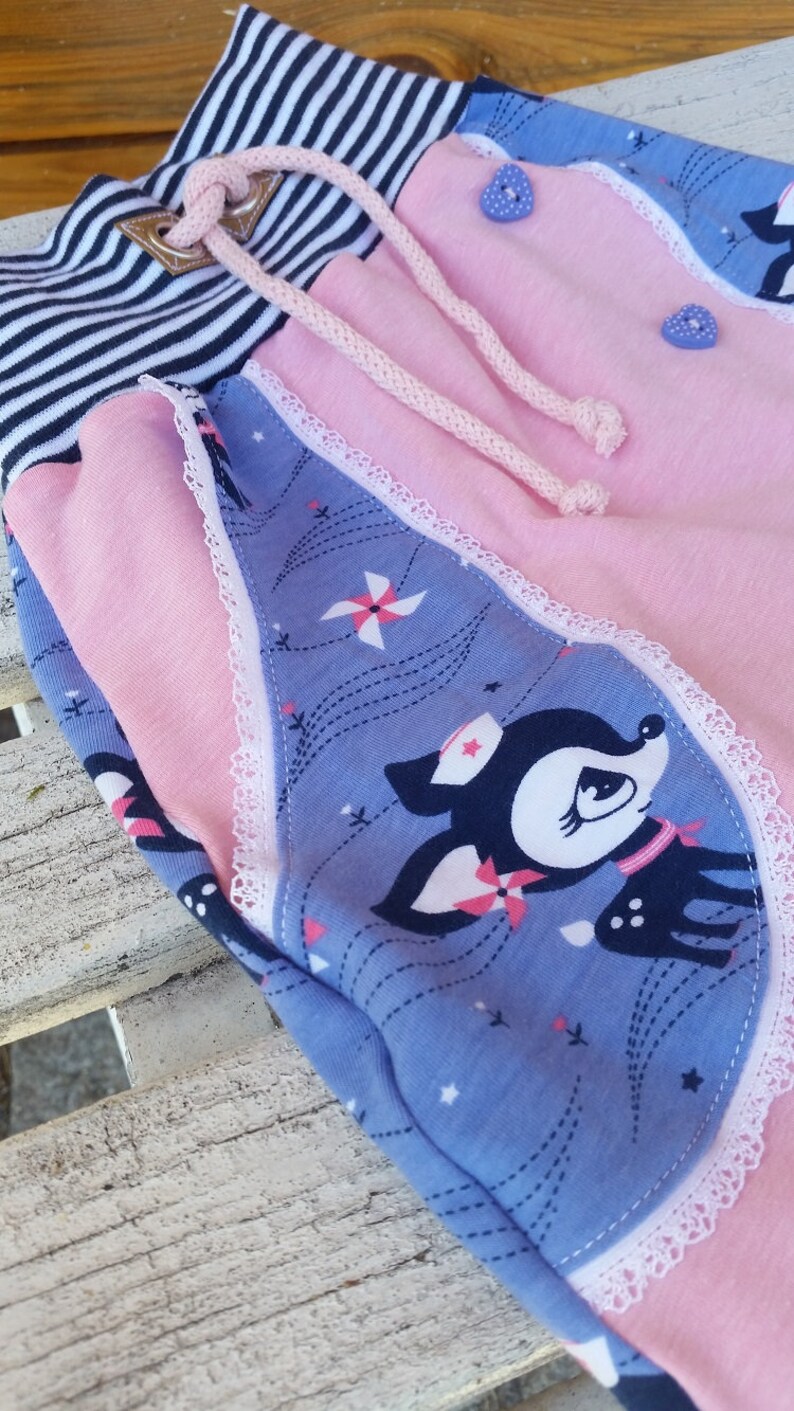K&ouml;nnte beinhalten: Rosa und blaue Kinderhose mit wei&szlig;er Spitzenborte und einem Hirschdruck. Die Hose hat einen gestreiften Bund mit einem Kordelzug.