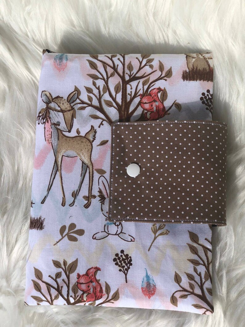 K&ouml;nnte beinhalten: Eine wei&szlig;e Wickelunterlage mit einer braunen und wei&szlig;en gepunkteten Tasche. Die Unterlage zeigt einen Wald-Druck mit Hirschen, Eichh&ouml;rnchen und Hasen.
