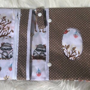 K&ouml;nnte beinhalten: Eine braune und wei&szlig;e Stoff-Wickeltasche mit Rei&szlig;verschluss, zwei Druckkn&ouml;pfen und einer Klappe mit einem Ausschnitt f&uuml;r ein Taschentuch. Der Stoff zeigt einen Waldtier-Druck mit Waschb&auml;ren, Eulen und F&uuml;chsen.