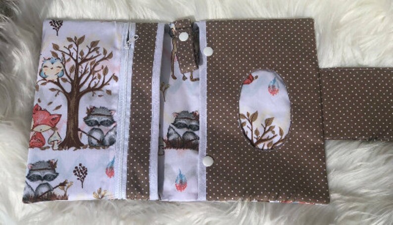 K&ouml;nnte beinhalten: Eine braune und wei&szlig;e Stoff-Wickeltasche mit Rei&szlig;verschluss, zwei Druckkn&ouml;pfen und einer Klappe mit einem Ausschnitt f&uuml;r ein Taschentuch. Der Stoff zeigt einen Waldtier-Druck mit Waschb&auml;ren, Eulen und F&uuml;chsen.