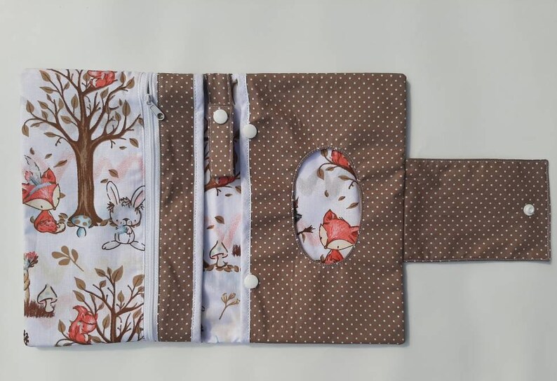 K&ouml;nnte beinhalten: Eine braune und wei&szlig;e Stoff-Wickeltasche mit Rei&szlig;verschluss, einer Klappe mit Druckknopfverschluss und einer Seitentasche. Der Stoff hat einen wei&szlig;en Hintergrund mit braunen Punkten und einen wei&szlig;en Hintergrund mit einem Waldtier-Druck. Die Tasche hat einen Taschentuchhalter mit einer ovalen Aussparung.