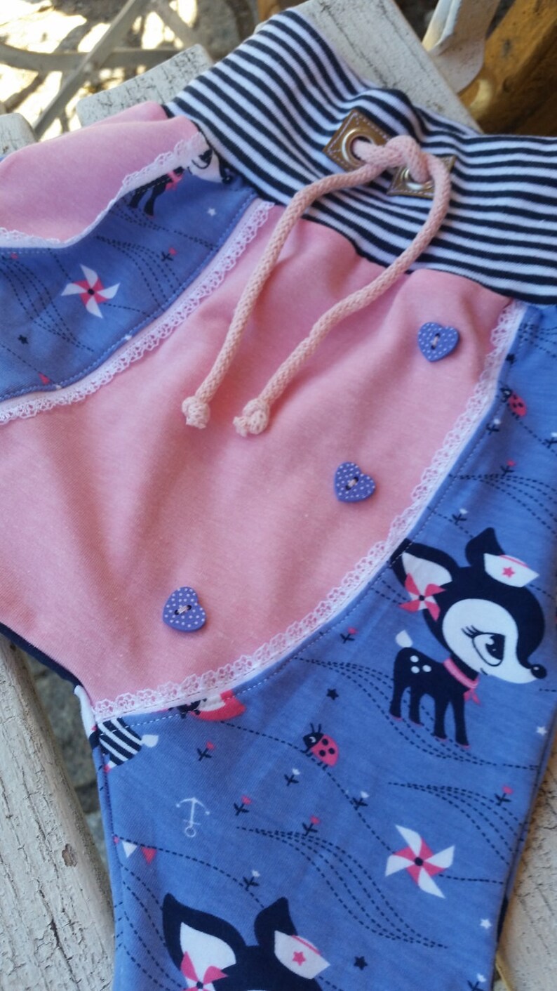 K&ouml;nnte beinhalten: Rosa und blaue Stoffhose mit wei&szlig;er Spitzenborte, einem schwarz-wei&szlig; gestreiften Bund und einem Kordelzug. Die Hose hat ein verspieltes Design mit Rehen, Marienk&auml;fern und Windm&uuml;hlen.