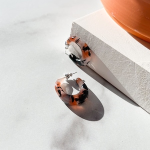 May include: A pair of hoop earrings with a tortoise shell pattern. The earrings are made of a translucent plastic material with black and brown swirls. The earrings are on a white surface.