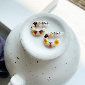 May include: A pair of small hoop earrings with a checkered pattern in brown, white, and yellow. The earrings have silver posts.