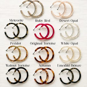 May include: A collection of twelve pairs of hoop earrings in various colors and patterns, labeled as Ghost, Cottage Core Pink, Meteorite, Ruby Red, Desert Opal, Peridot, Original Tortoise, White Opal, Walnut Tortoise, Autumn, and Emerald Bronze.