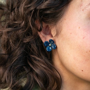 Puede incluir: Un par de pendientes de color azul en forma de flor con detalles dorados. Los pendientes los lleva una persona con el pelo oscuro y rizado.