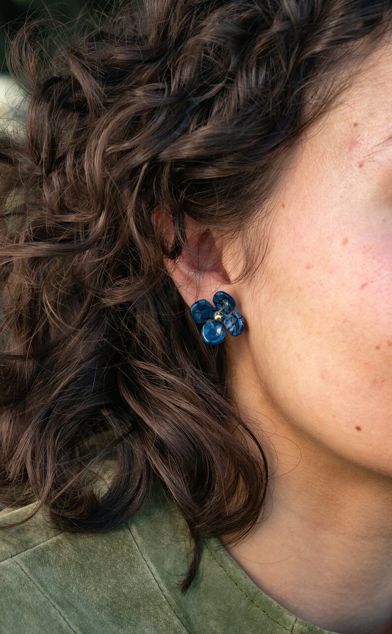 Puede incluir: Un par de pendientes de color azul en forma de flor con detalles dorados. Los pendientes los lleva una persona con el pelo oscuro y rizado.