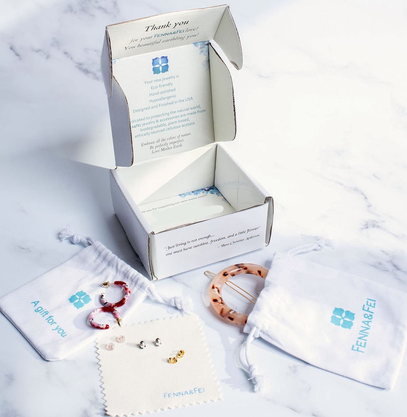 Puede incluir: Una caja blanca con el texto "&iexcl;Gracias por tu amor FENNA&FEI!" y un dise&ntilde;o de flor azul. La caja est&aacute; abierta y contiene una bolsa blanca con el texto "Un regalo para ti" y un pendiente con estampado rojo y blanco en su interior. Tambi&eacute;n hay una bolsa blanca con el texto "FENNA&FEI" y una pinza para el pelo marr&oacute;n y blanca en su interior. La caja tambi&eacute;n contiene una tarjeta blanca con el texto "FENNA&FEI" y tres pares de pendientes.
