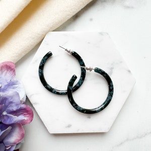 May include: A pair of circular hoop earrings featuring a dark black and teal speckled pattern. The earrings are constructed with a smooth, polished finish and include silver-toned metal posts and backings for secure attachment to the earlobes.