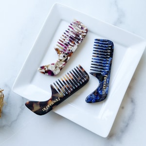 Könnte beinhalten: Drei Schildpattkämme mit einem Griff auf einem weißen Teller. Die Kämme sind aus einem durchscheinenden Material mit einem Marmormuster gefertigt. Die Griffe sind mit dem Wort "FENNABELLE" in goldenen Buchstaben verziert.