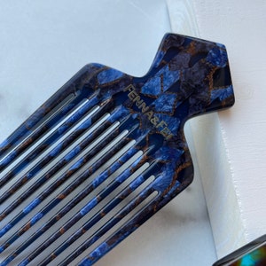 K&ouml;nnte beinhalten: Ein blauer und goldener Kamm mit einem einzigartigen Design. Der Kamm hat einen breiten Griff mit dem aufgedruckten Text "FENNA & FEI".