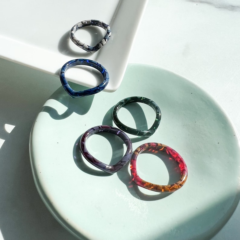 May include: Five colorful rings made of resin with different patterns. The rings are on a light blue surface.