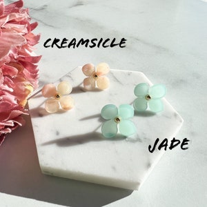 May include: Two pairs of flower-shaped earrings. One pair is a light pink colour with the text "CREAMSICLE" above them. The other pair is a light blue colour with the text "JADE" below them.