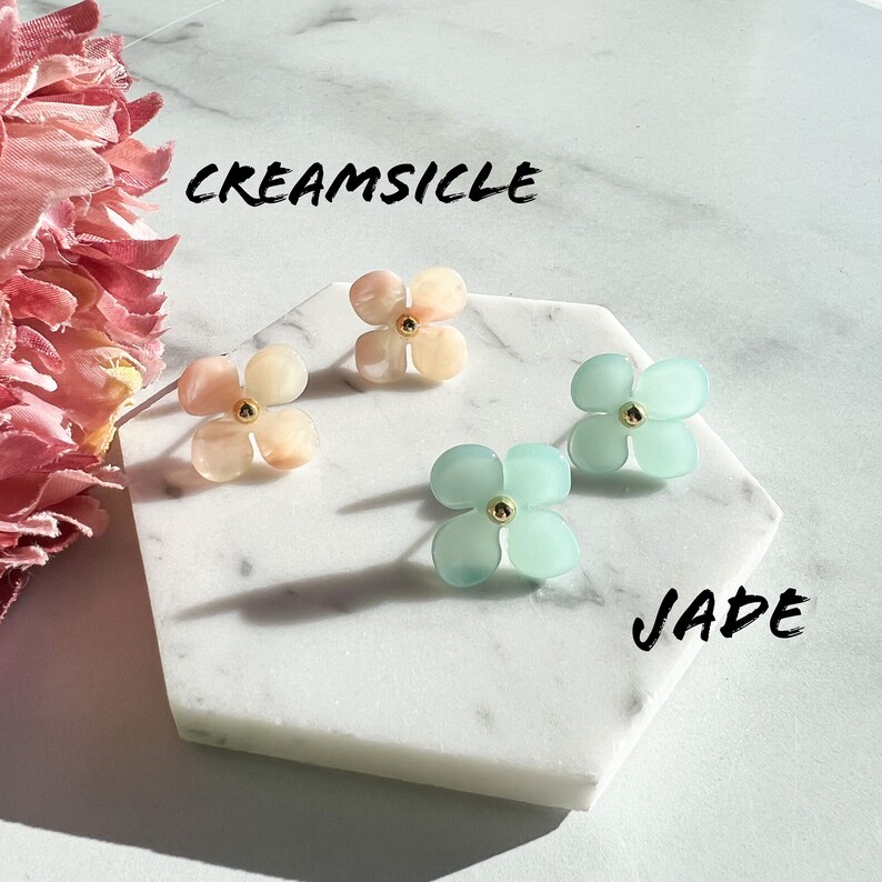 May include: Two pairs of flower-shaped earrings. One pair is a light pink colour with the text "CREAMSICLE" above them. The other pair is a light blue colour with the text "JADE" below them.