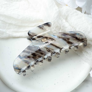 May include: A marbled, claw-style hair clip in shades of white, gray, and brown. The clip features a gold-colored emblem and the text "FENNA&FEI". The hair accessory is designed with a strong grip and is suitable for various hair types.
