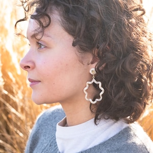 Puede incluir: Una mujer con el pelo rizado casta&ntilde;o lleva un c&aacute;rdigan gris y una camiseta blanca. Lleva unos pendientes grandes de estrella blanca.