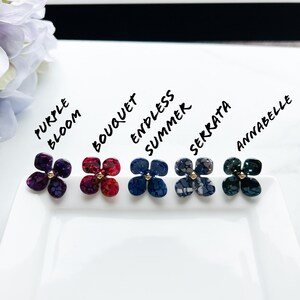 May include: Six different coloured flower-shaped earrings with gold centres. The earrings are labelled "Purple Bloom", "Bouquet", "Endless Summer", "Serrata", "Annabelle".