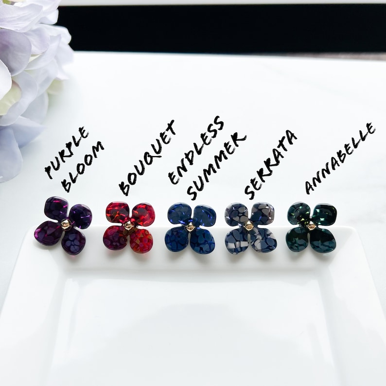 May include: Six different coloured flower-shaped earrings with gold centres. The earrings are labelled "Purple Bloom", "Bouquet", "Endless Summer", "Serrata", "Annabelle".