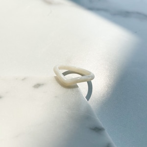May include: A white, opaque, ring with a unique, slightly curved, rectangular shape. The ring is sitting on a white marble surface.