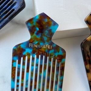 K&ouml;nnte beinhalten: Ein blauer und brauner Schildpattkamm mit einem breiten Zahn-Design. Der Kamm hat eine einzigartige Form mit einer spitzen Oberseite und einer breiten Basis. Der Kamm ist aus Kunststoff und hat die Aufschrift "Fenna & Fei" auf der Vorderseite.