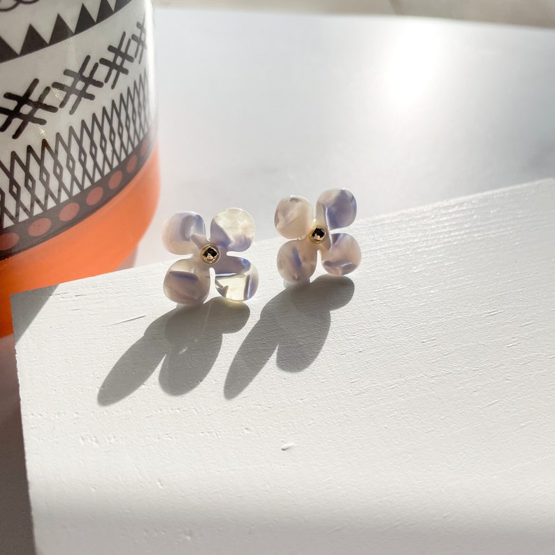 Puede incluir: Un par de peque&ntilde;os pendientes en forma de flor blanca con un patr&oacute;n de espiral de color p&uacute;rpura claro y un centro dorado.
