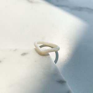 以下が含まれることがあります： 白く不透明なリングで、ユニークなわずかに湾曲した長方形の形状をしています。リングは滑らかで磨かれた素材で作られており、白い大理石の表面の上に置かれています。