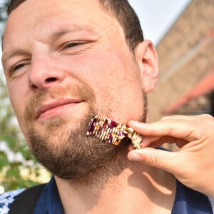 Puede incluir: Un peine para barba peque&ntilde;o y rectangular con un patr&oacute;n jaspeado en tonos crema, marr&oacute;n y morado oscuro. El peine tiene una serie de dientes finos y muy juntos a lo largo de un borde y una secci&oacute;n de mango curva para sujetarlo.