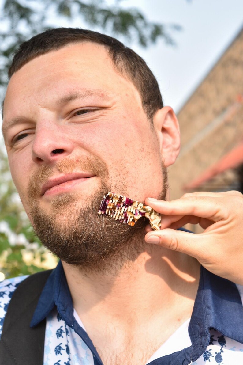 Puede incluir: Un peine para barba peque&ntilde;o y rectangular con un patr&oacute;n jaspeado en tonos crema, marr&oacute;n y morado oscuro. El peine tiene una serie de dientes finos y muy juntos a lo largo de un borde y una secci&oacute;n de mango curva para sujetarlo.