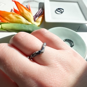 May include: A gray and black patterned ring with a unique V-shaped design. The ring is worn on a person's finger.