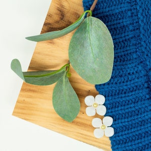 Puede incluir: Dos pendientes blancos en forma de flor con centros dorados sobre una superficie de madera con hojas verdes y una tela azul de punto.