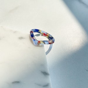 以下が含まれることがあります： 樹脂製のカラフルな幾何学模様のリング。リングは角が丸みを帯びた正方形で、青、赤、黄、白のパターンが施されています。