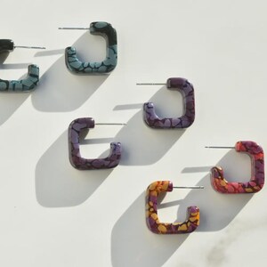 May include: Five square-shaped hoop earrings featuring a multi-colored mosaic pattern. The earrings are arranged in a scattered layout and include shades of blue, purple, and orange, each with a silver-toned metal post for pierced ears.