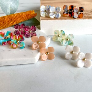 Puede incluir: Surtido de pendientes con forma de flor de colores con detalles dorados. Los pendientes est&aacute;n hechos de varios materiales, incluyendo pl&aacute;stico, resina y concha.